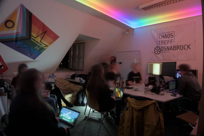 Ein Foto vom Hackspace. Um einen Tisch und auf einem Sofa sitzen ein paar Menschen mit Laptops. An den Wänden hängt ein Banner mit dem Chaostreff-Osnabrück-Logo, eine Progress-Flagge und ein Whiteboard. Die Beleuchtung ist ein LED-Streifen mit Regenbogenfarben.